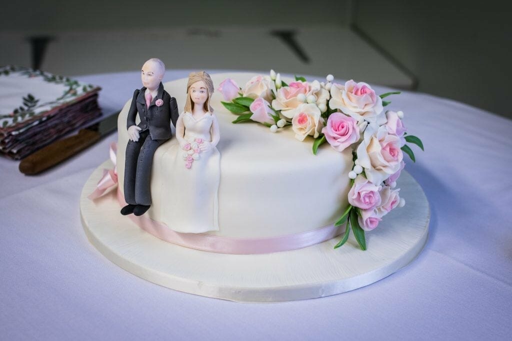 A wedding cake depicting the bride and groom sitting on top next to some pink and white flowers.